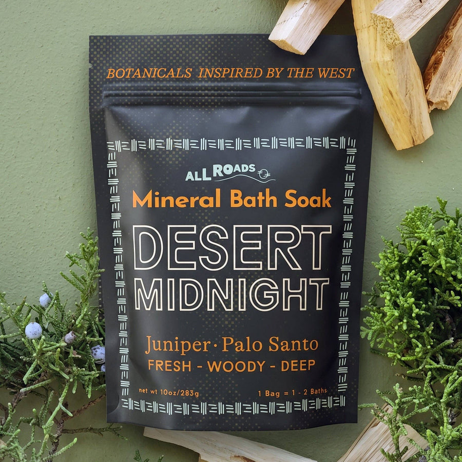 A black resealable pouch of All Roads Mineral Bath Soak, Desert Midnight, styled on a sage green background with natural wood and greenery props.