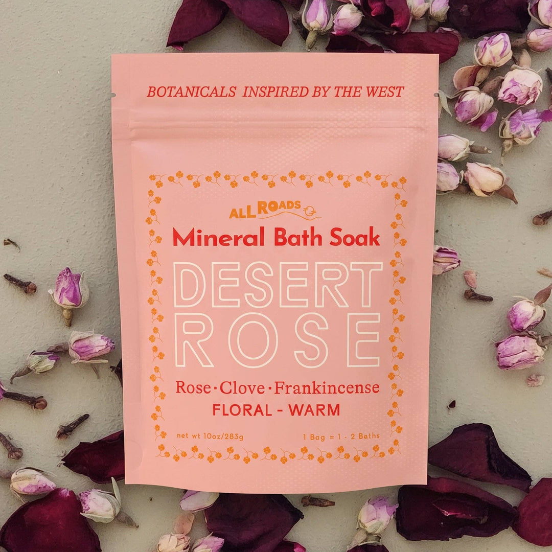 A pink stand-up pouch of All Roads Mineral Bath Soak in Desert Rose scent, surrounded by scattered dark rose petals and small flower buds on a light neutral surface.